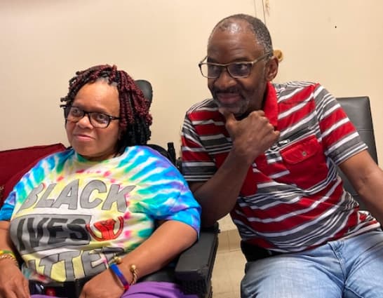 Una foto de una persona de pelo negro con una camisa teñida de corbata que dice Black Lives Matter (Las vidas de los negros importan) se sienta junto a una persona con gafas que lleva un polo a rayas.