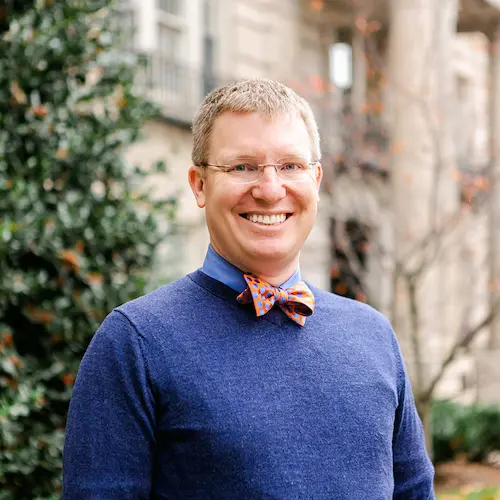 Headshot of John Stowe. John wears a blue sweater and a bow tie.