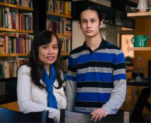 A photo of two people beside each other and looking at the camera. On the left is a seated woman in a white sweater, and on the right is a young man in a striped shirt. 
