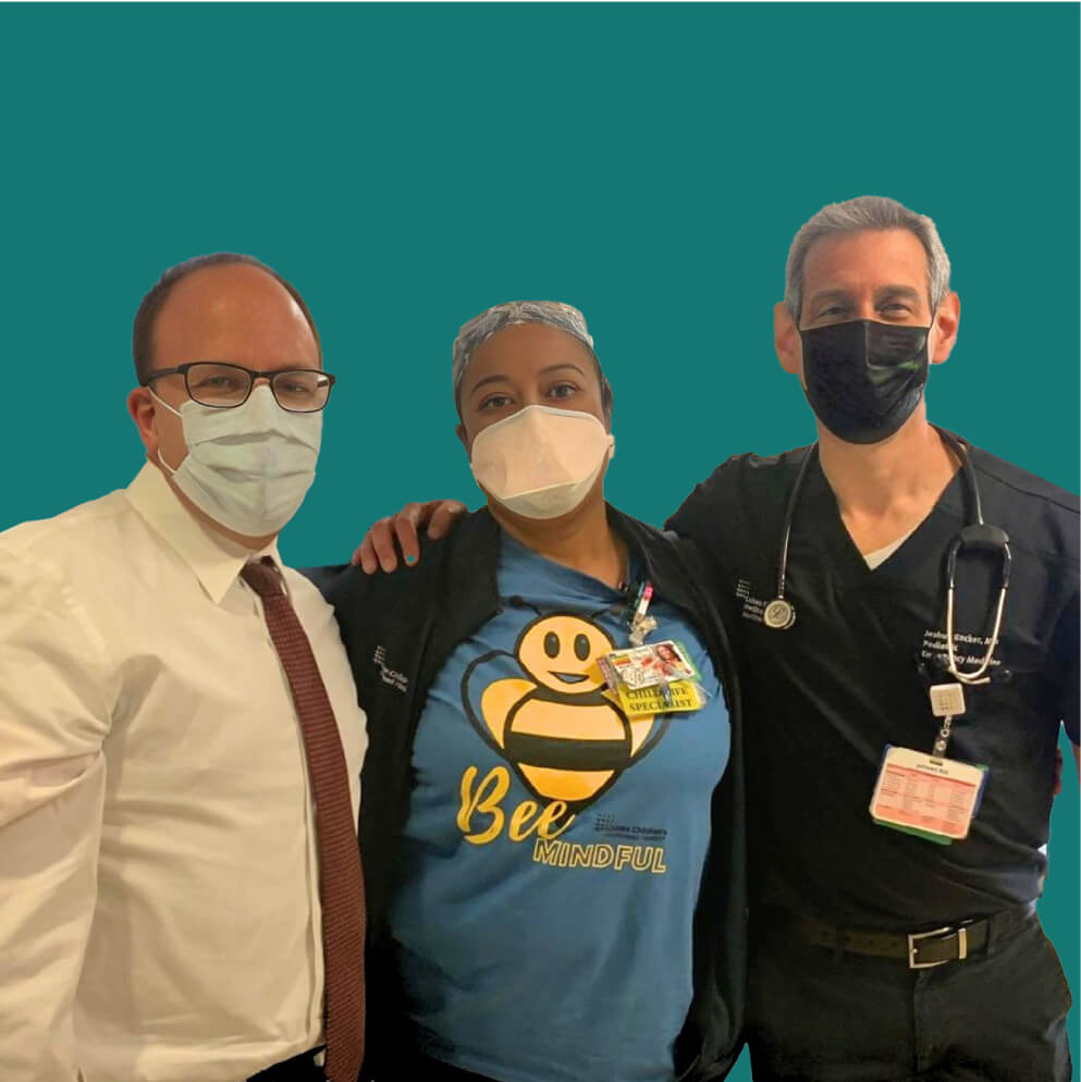 Foto de tres personas con mascarillas que miran a la cámara. La persona de la izquierda lleva una camisa blanca, la persona del medio lleva una camisa azul y la persona de la derecha lleva una bata negra y un estetoscopio.