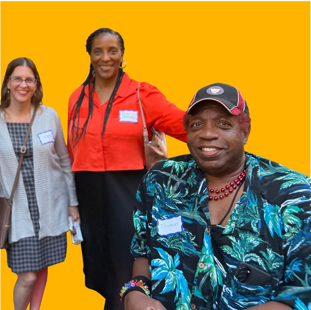 A photo of three people smiling and looking at the camera. The woman on the left wears a dress with a white cardigan, a woman in the middle has long hair and wears an orange shirt, and a person on the right who is seated and wears a Hawaiian shirt.