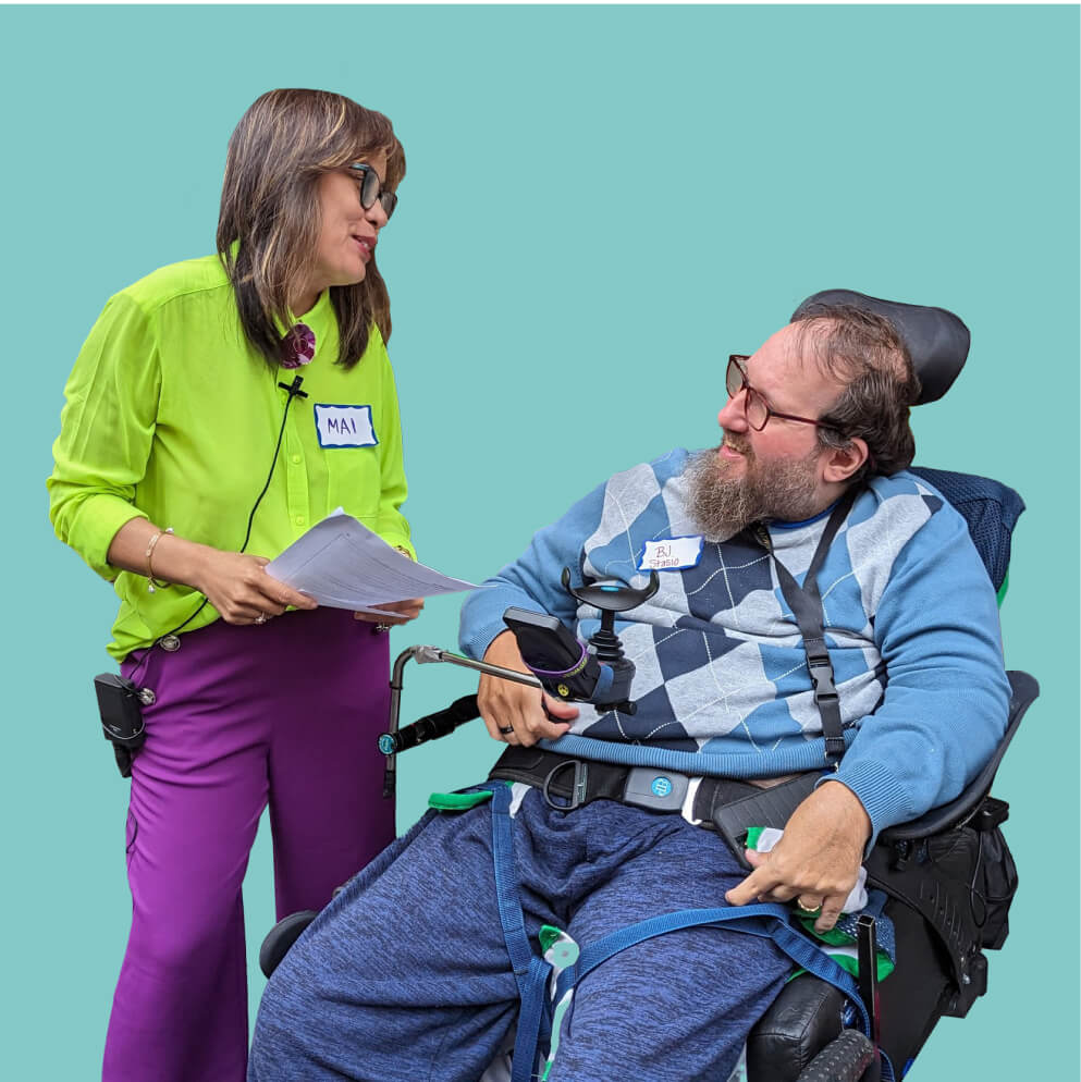 A photo of two people smiling and looking at each other. The person on the right wears a green shirt and is standing. The person on the right has a beard and glasses and uses a wheelchair.