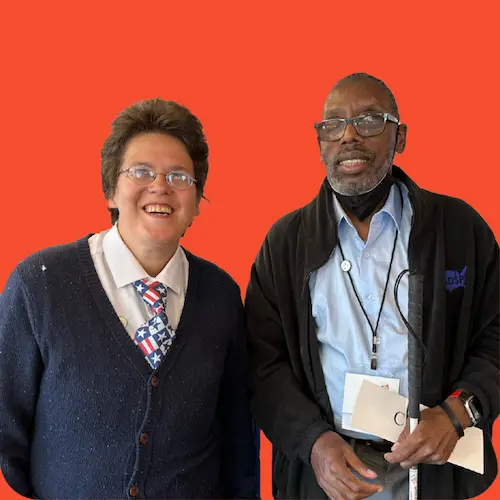 Una foto de dos personas de pie una junto a la otra y sonriendo a la cámara. La persona de la izquierda lleva el pelo corto y gafas, y la de la derecha es calva y lleva gafas.